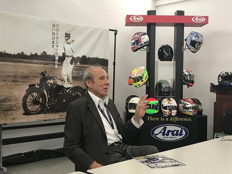 At 85 years old, Michio Arai reflects on Arai Helmet's rich history in Saitama. Behind him, an iconic photograph captures his father and the company's founder, Hirotake, standing on the saddle of his Harley in the late 1930s, embodying the enduring spirit and legacy of the Arai family in the world of motorcycle helmets.