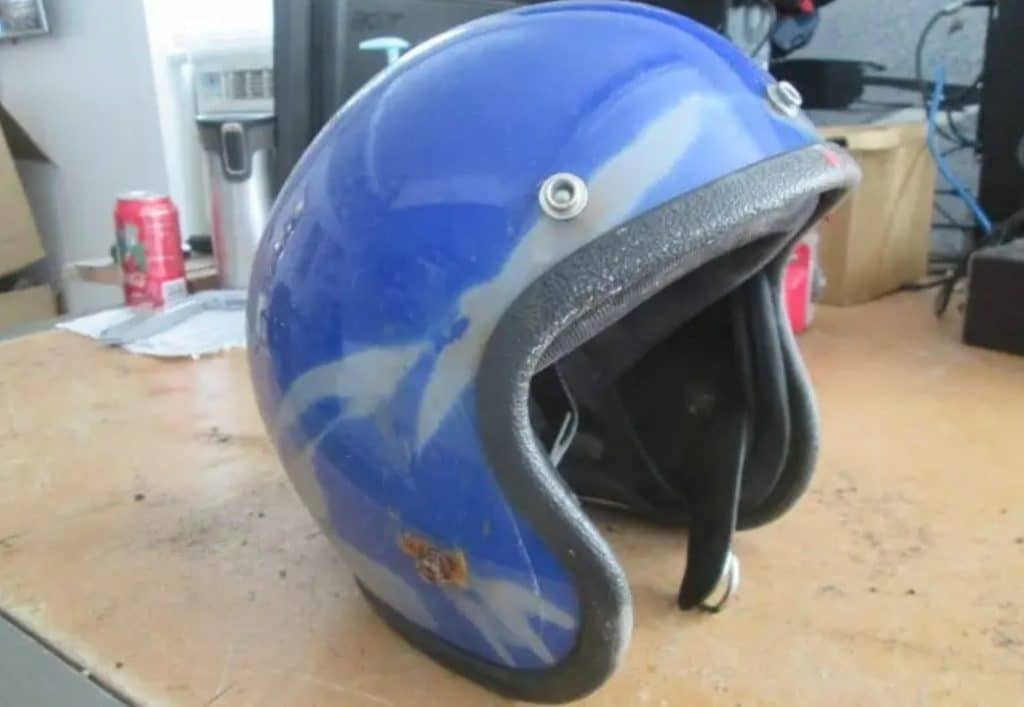 The worn-out dark blue AGV Valenza open face helmet exhibits clear signs of sun exposure damage, with streaks of faded paint and rusty metal appendages. Direct sunlight and exposure to humidity not only deteriorate the paintwork but also weaken the structural integrity of the helmet.