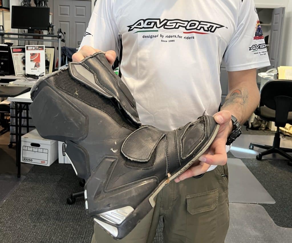 A well-worn 2005 AGVSPORT Laguna race boot, displayed by Denis Grachev, a renowned stunt rider and owner of OFF LIMITS TOURS, exhibits visible signs of a crash and wear in high-friction areas. Despite nearly 20 years of use, this boot demonstrates its durability, having effectively protected the rider last year during a high-speed crash exceeding 80 mph.